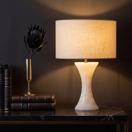The Luxury Concave Table Lamp with a cream shade glows on a dark marble surface beside stacked books and a gold vase with a dried flower, all set against a deep-toned wall.