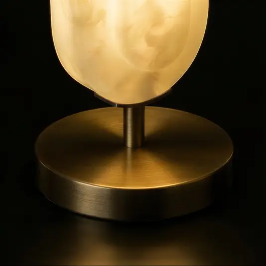 A close-up of the Opal Elipse Step Table Lamp, showcasing its round brushed brass base and softly glowing marble-patterned glass shade against a dark backdrop.