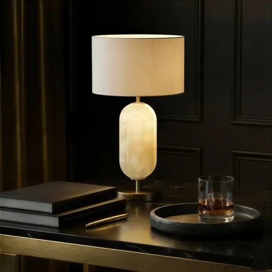 An Opal Ellipse Step Table Lamp with a white cylindrical shade and oval base rests on a black marble desk beside a tray holding whisky, a pen, and stacked black notebooks. The setup is set against dark panelled walls.
