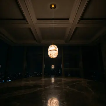 Luxury Banded Stone Ellipse Light A single Banded Elipse Pendant Light – Antique Brass hangs from the coffered ceiling, casting a warm glow on the polished marble, as city lights shimmer beyond the dark windows in the background.