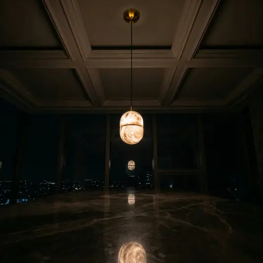 A single Banded Elipse Pendant Light – Antique Brass hangs from the coffered ceiling, casting a warm glow on the polished marble, as city lights shimmer beyond the dark windows in the background.