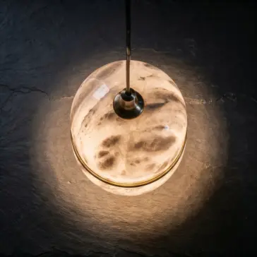 Luxury Banded Stone Ellipse Light The Banded Ellipse Pendant Light – Antique Brass hangs from a black flex, its round, banded design casting a warm, ambient glow over the dark textured surface below.