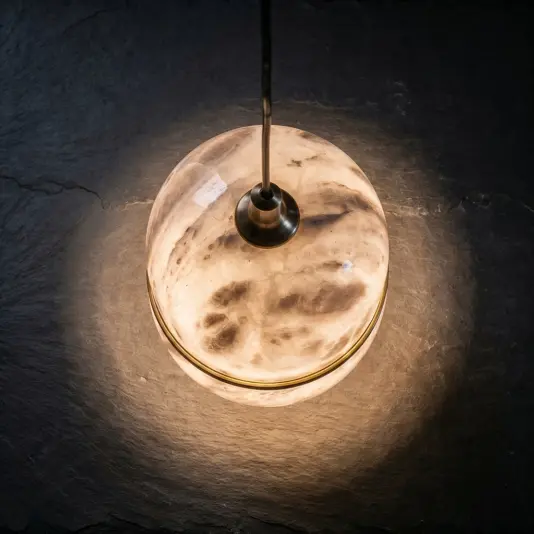 The Banded Ellipse Pendant Light – Antique Brass hangs from a black flex, its round, banded design casting a warm, ambient glow over the dark textured surface below.