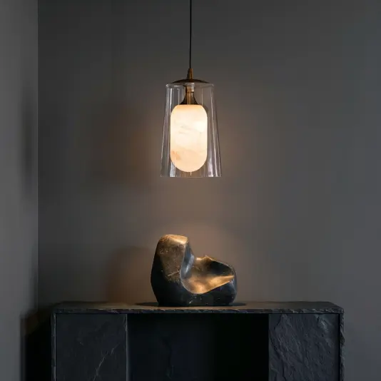 An Elegant Alabaster Glass Tapered Pendant Light with a frosted bulb and glass shade hangs above a dark stone sculpture on a slate shelf set against a grey wall.