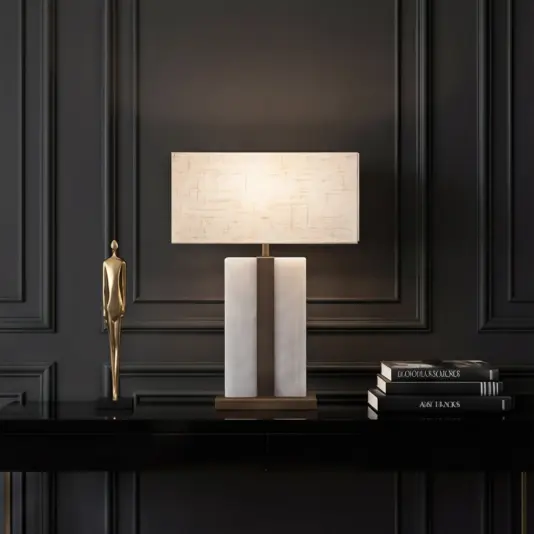 A modern table lamp with a rectangular white shade sits on a black console table, next to a gold human figurine and stacked books, against an elegant dark panelled wall.