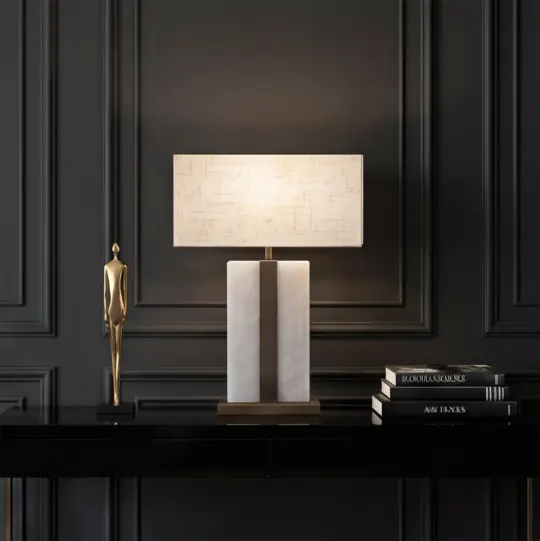 A modern table lamp with a rectangular white shade sits on a black console table, next to a gold human figurine and stacked books, against an elegant dark panelled wall.