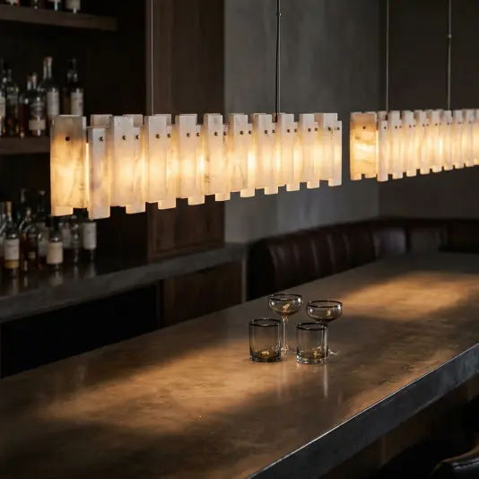 Linear Stone Panel Bar Pendant Light A modern bar features a long, softly glowing Linear Alabaster Stone Panel Bar Pendant above a polished counter. Three smoky glass tumblers and one coupe glass rest on the counter, with shelves of liquor bottles in the background.