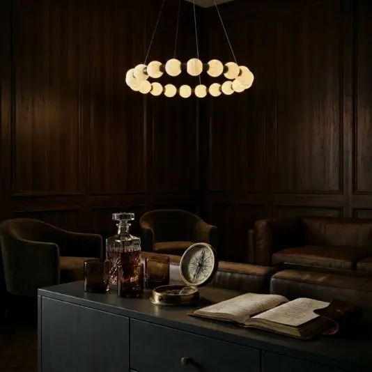 A dimly lit, elegant room with dark wood panelling and leather chairs glows softly under a Luxury Alabaster Sphere Ring Pendant – Circular Stone Chandelier. On the table: a decanter, glass, compass, magnifying glass, and an open book.