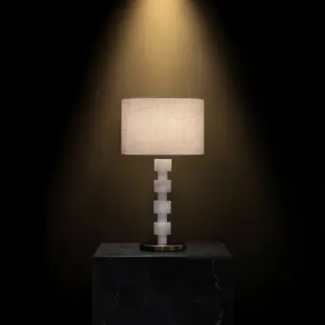 The Opal Round Stacked Table Lamp has a textured beige shade and a geometric stacked white base on a dark marble plinth, showcased under focused light against a black background.