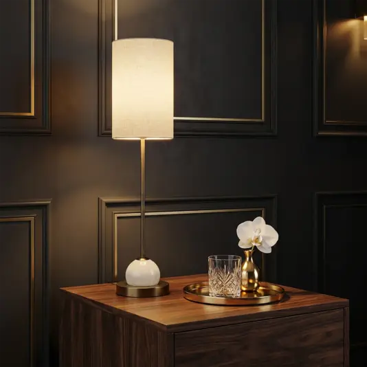 The Luxury Tall Globe with Rod Table Lamp with a beige shade is placed on a wooden bedside table beside a gold tray that holds a glass tumbler, crystal glass, and a white orchid in a gold vase against a dark panelled wall.