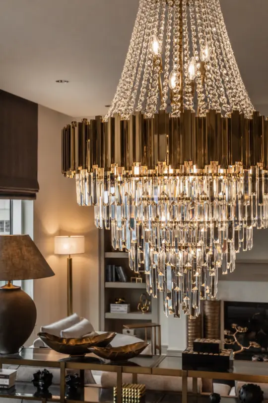 A luxurious living room features a large, elegant crystal chandelier with cascading prisms, modern decor, a table lamp, decorative objects, bookshelves, and a fireplace in the background.