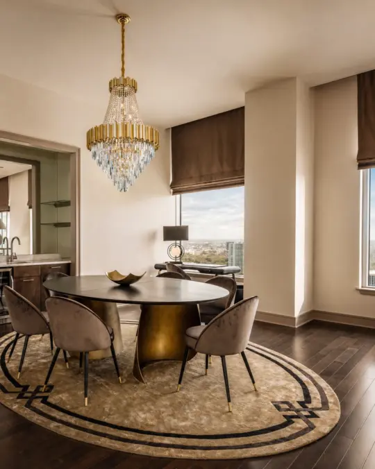 A modern dining room with a round table, six upholstered chairs, a gold and crystal chandelier, large windows with brown blinds, a beige patterned rug, and dark hardwood flooring. A city view is visible outside.