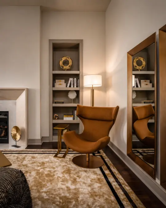 A cosy modern living room corner with a brown swivel chair, gold side table, floor lamp, large mirror, built-in shelves with décor, and a beige rug on dark wood flooring.