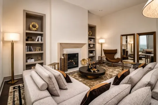 A modern living room with a beige corner sofa, brown accent cushions, a round black coffee table, a fireplace, built-in shelves with décor, a brown lounge chair, standard lamps, and two tall mirrors reflecting a city view.