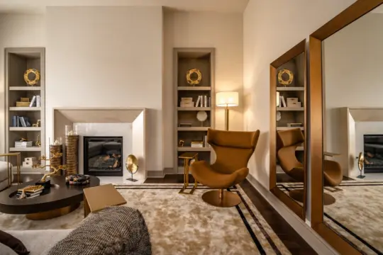 A modern living room with a brown armchair, large gold-framed mirror, built-in shelves with gold decor, a fireplace, round coffee table, floor lamp, and neutral tones throughout.