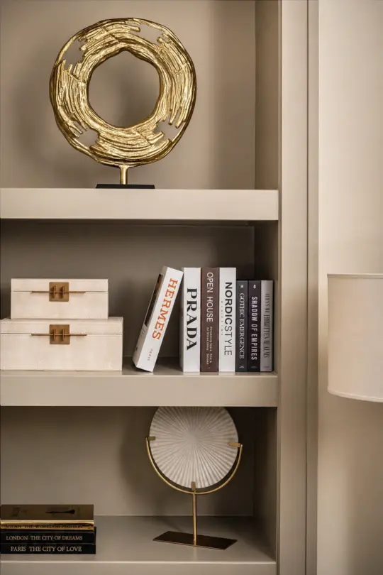 A beige shelving unit displays decorative objects, books, two cream-coloured boxes with gold clasps, a gold circular sculpture, a white fan-shaped ornament on a stand, and stacked books about major cities.