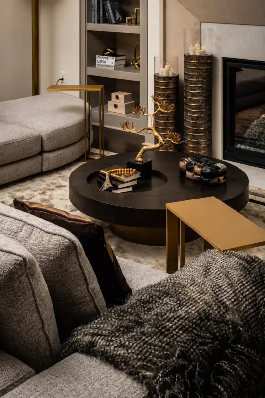 Modern living room with beige sofas, a round black coffee table with decorative items, gold accent tables, candles, a cosy throw blanket, and a built-in shelf beside a fireplace. Warm, elegant, and inviting atmosphere.