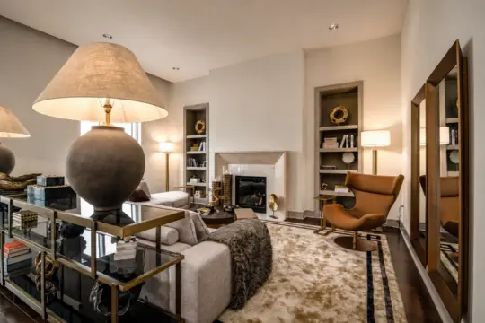 A modern living room with neutral tones features a large lamp, cosy sofa, brown armchair, fireplace, bookcases, tall mirror, and decorative accents on shelves, all set on a plush beige rug.