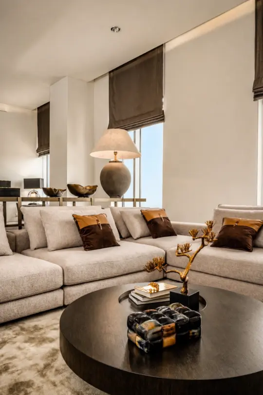 Modern living room with a beige corner sofa, brown accent cushions, a round dark coffee table with decorative items, a large lamp, and floor-to-ceiling windows with brown Roman blinds. Neutral, cosy, and elegant decor.