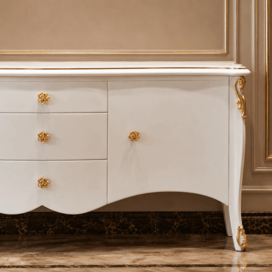 Large Classic Style Sideboard Elegant white chest of drawers with gold accents and ornate gold knobs, set against a beige wall with gold trim, standing on a marble floor.