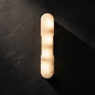 Modern lozenge shape Wall Light A modern, elongated wall sconce with rounded ends emits a warm glow against a dark textured wall, casting soft light and creating gentle shadows.
