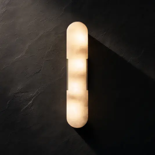 A modern, elongated wall sconce with rounded ends emits a warm glow against a dark textured wall, casting soft light and creating gentle shadows.