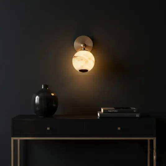 Modern Orb Wall Light A round marble wall sconce emits warm light above a black console table with gold legs, a black vase, and stacked books, all set against a dark wall.