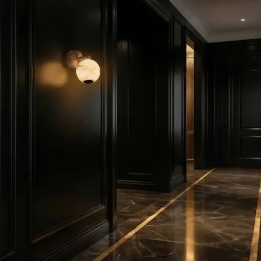 Modern Orb Wall Light A modern hallway with glossy dark walls and a marble floor features a single lit wall light with a round, marbled glass shade, casting a warm glow. The space feels elegant and luxurious, with gold trim on the floor.