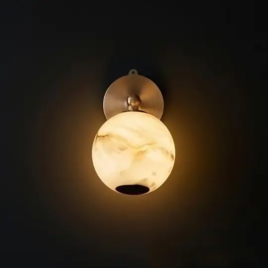 A round wall sconce with a marble-patterned glass shade emits a warm, soft light against a dark background. The fitting has a metallic, circular base mounted on the wall.