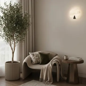 Modern Alabaster Round Wall Light A cosy reading nook features a curved chair, green cushion, beige throw, and a small table with books and a cup. An indoor tree and the Modern Round Wall Light enhance the inviting atmosphere of this neutral-toned space.