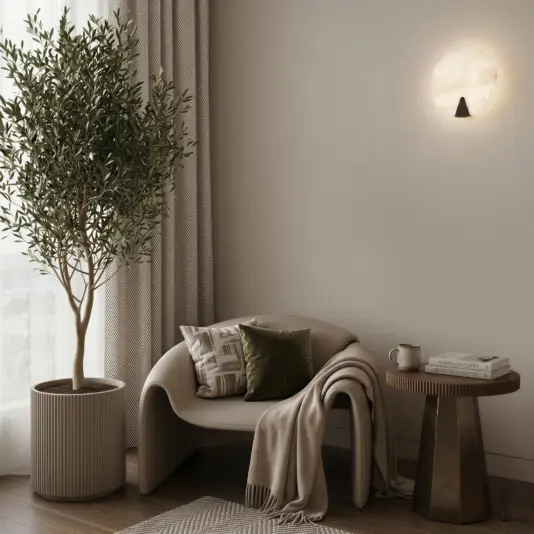 Modern Alabaster Round Wall Light A cosy reading nook features a curved chair, green cushion, beige throw, and a small table with books and a cup. An indoor tree and the Modern Round Wall Light enhance the inviting atmosphere of this neutral-toned space.