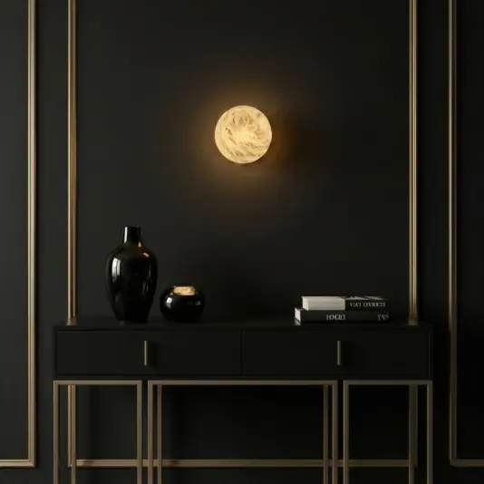 Luxury Round Wall Light A modern black console table with gold trim holds black vases and stacked books, set against a dark wall with gold accents and a round, glowing, marble-like wall sconce above it.