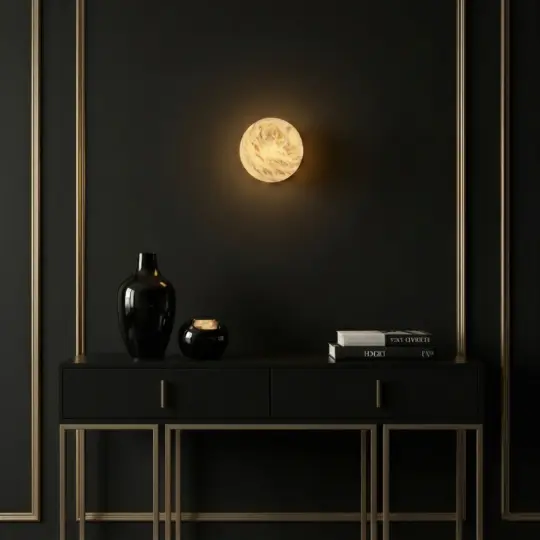A modern black console table with gold trim holds black vases and stacked books, set against a dark wall with gold accents and a round, glowing, marble-like wall sconce above it.