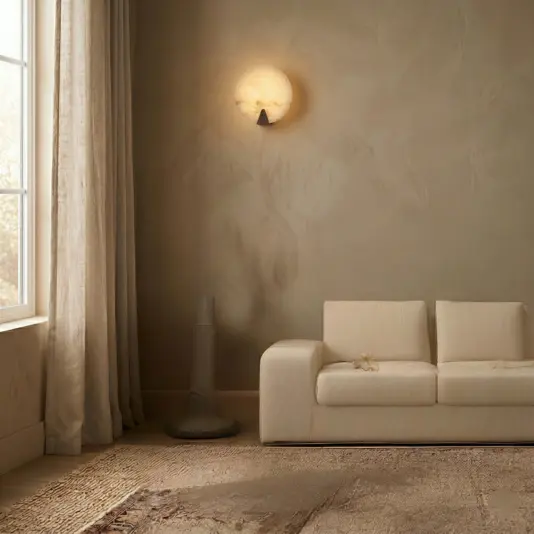 Modern Alabaster Round Wall Light A minimal living room with an off-white sofa, textured beige wall, Modern Round Wall Light, floor-length curtains, tall fan or speaker, and a large neutral-toned rug. Sunlight streams in from a window on the left.