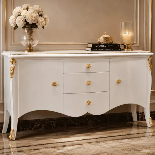 Elegant white sideboard with gold accents, featuring ornate drawer handles and legs. On top are a glass vase with white flowers, stacked books, a decorative box, and a glass candle holder. Marble floor and panelled wall background.