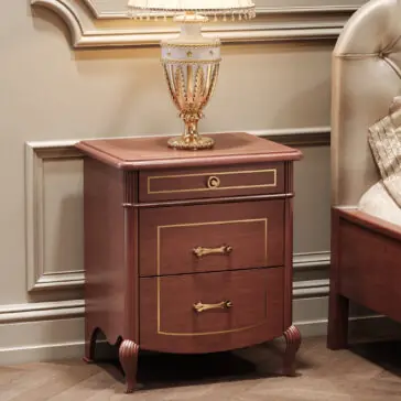 The Classic Style Briarwood Bedside Cabinet with gold accents and ornate handles on two drawers stands beside a tufted headboard bed, topped with a decorative gold and white lamp in an elegant room.