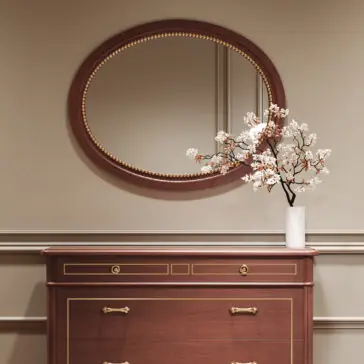 Classic Style Cherrywood Oval Beaded Mirror A Classic Style Cherrywood Chest of Drawers (Copy) with gold accents stands against a beige wall, topped with a white vase of flowering branches. Above, an oval mirror with a wooden and gold-trimmed frame reflects the room.