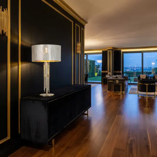 Elegant living room with dark walls and gold trim, featuring the Gold Bamboo Style Table Lamp on a black sideboard, wooden floors, chic furniture, and large windows framing a scenic evening city view.