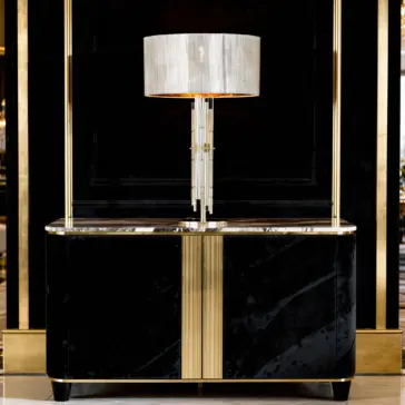 A modern black and gold sideboard with a glossy finish stands against a dark wall, topped with a tall, elegant lamp featuring a cylindrical white shade and vertical glass rods.