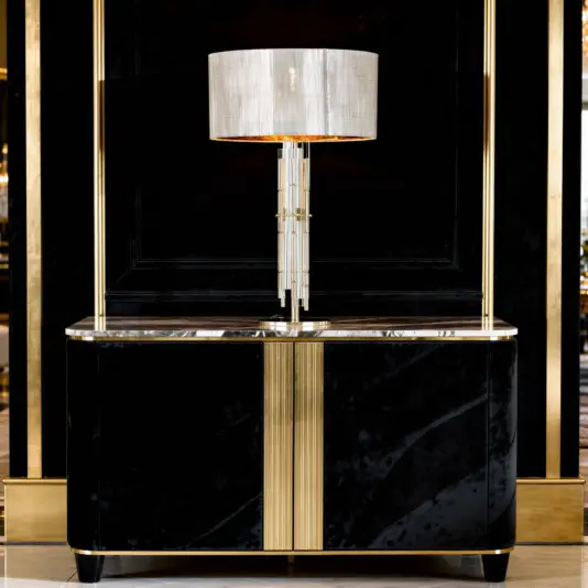 A modern black and gold sideboard with a glossy finish stands against a dark wall, topped with a tall, elegant lamp featuring a cylindrical white shade and vertical glass rods.