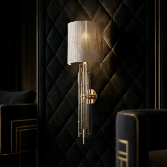 A modern wall sconce with a cylindrical fabric shade and cascading metallic rods is mounted on a black, quilted wall in a luxurious, dimly lit room featuring dark upholstered furniture with gold accents.