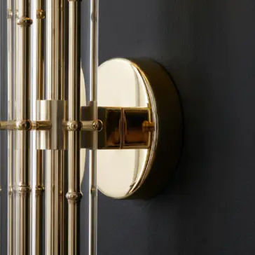 Close-up of a modern wall sconce featuring a round, polished gold base and multiple vertical gold and glass rods, mounted on a dark, textured wall.