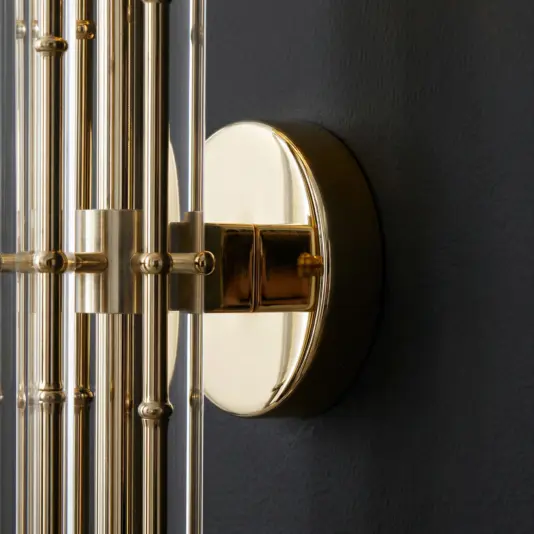 Close-up of a modern wall sconce featuring a round, polished gold base and multiple vertical gold and glass rods, mounted on a dark, textured wall.