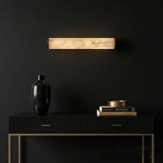 A modern, minimalist black console table with gold accents holds a black vase and a stack of books with a gold bowl on top. Above, a wood-textured wall sconce emits warm light against a dark wall.