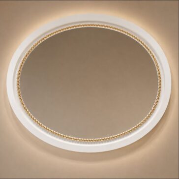 An oval mirror with a white and gold beaded frame is mounted on a beige wall, illuminated softly from behind, creating a warm and elegant appearance.