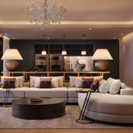 A modern living room with a large beige corner sofa, decorative cushions, round coffee table, two table lamps, wooden floors, built-in bookshelves, a chandelier, and an open kitchen in the background.