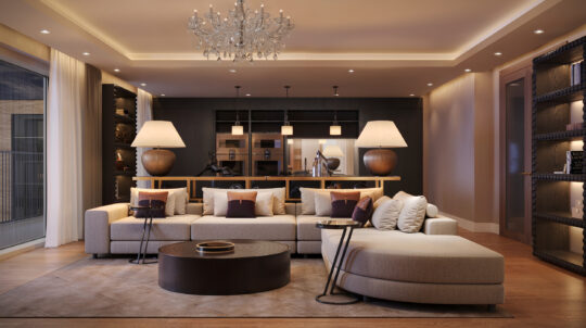 A modern living room with a large beige corner sofa, decorative cushions, round coffee table, two table lamps, wooden floors, built-in bookshelves, a chandelier, and an open kitchen in the background.
