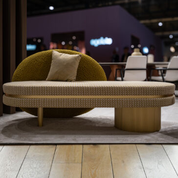 The Contemporary Geometric Bench features a modern beige and olive-green design with a textured, rounded backrest, cylindrical base, and single beige cushion, set on a light rug over wooden flooring in an indoor space.