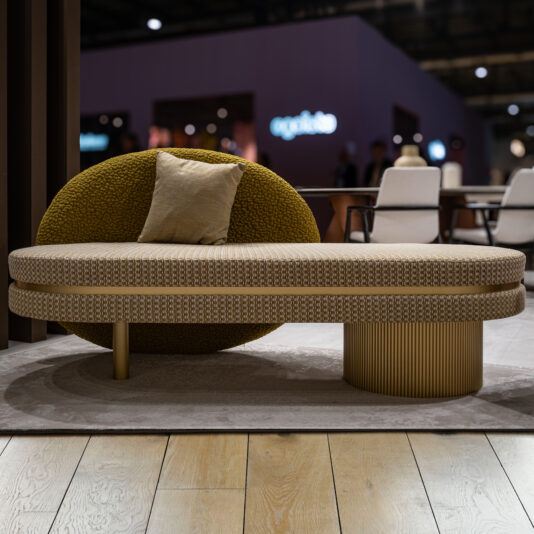 The Contemporary Geometric Bench features a modern beige and olive-green design with a textured, rounded backrest, cylindrical base, and single beige cushion, set on a light rug over wooden flooring in an indoor space.