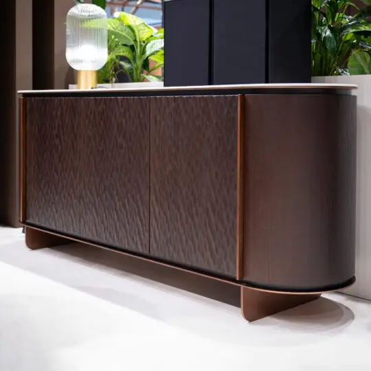 The Contemporary Hammered Oak Veneer Sideboard features textured doors, rounded edges, and a white tabletop. Style it with a glass lamp and green plants on a light floor for a modern indoor look.